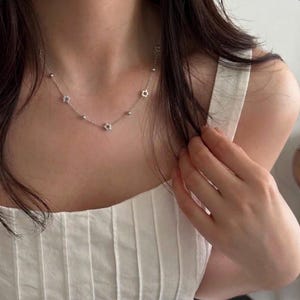 May include: A silver-toned chain necklace featuring small, evenly spaced metallic flower charms and tiny spherical beads. The delicate link chain rests against the neck, showcasing a repeating pattern of floral elements and smooth round accents.