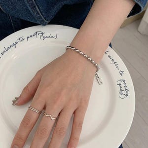 May include: A silver-toned beaded chain bracelet with a small rectangular tag and two silver-toned rings, one featuring a smooth band and the other a wavy design, worn on a wrist resting against a white plate with black cursive text.