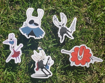 Puede incluir: Cinco pegatinas troqueladas con personajes de un videojuego. Las pegatinas representan varios personajes en diferentes poses, con una paleta de colores blanco, negro, rojo y azul. Las pegatinas están dispuestas sobre hierba verde.