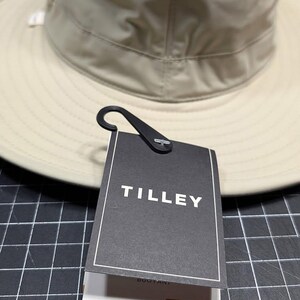 May include: A beige Tilley hat with a wide brim and a mesh crown for ventilation. The hat has a black tag with the brand name "TILLEY" printed on it. The tag also includes the "AIRFLO" and "UPF 50+" labels.