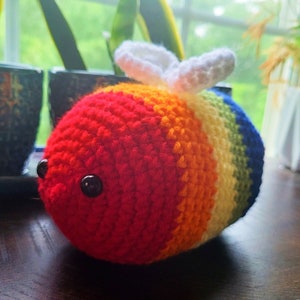 Puede incluir: Una abeja de ganchillo hecha a mano con colores del arcoíris. La abeja tiene rayas rojas, naranjas, amarillas, verdes y azules. Tiene ojos negros y alas blancas. Un objeto decorativo encantador.