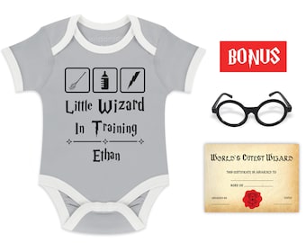 May include: A gray baby onesie with white trim features a black graphic of a broom, a baby bottle, and a lightning bolt. The text "Little Wizard In Training" is printed below the graphic, followed by the name "Ethan". The onesie is accompanied by a pair of black round glasses, a red rectangle with the word "BONUS" printed in white, and a certificate that reads "World's Cutest Wizard".