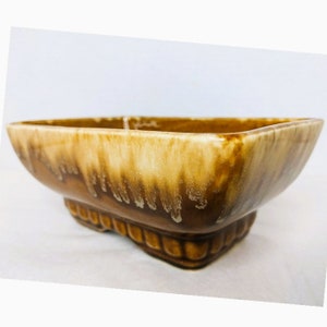May include: A brown and white glazed ceramic planter with a square shape and a raised base. The planter has a drip glaze effect.