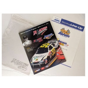 Può includere: Un programma souvenir per la gara NASCAR Talladega 500, con una macchina verde e gialla con il numero 55. La copertina del programma include anche il testo "Talladega 500" e "Aaron's" in rosso e bianco. Il programma è in una custodia di plastica trasparente.
