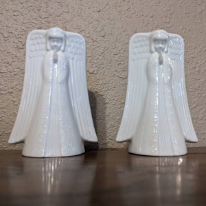 May include: Two white porcelain angel figurines with wings outstretched and hands clasped in prayer. The angels are standing on a wooden surface.