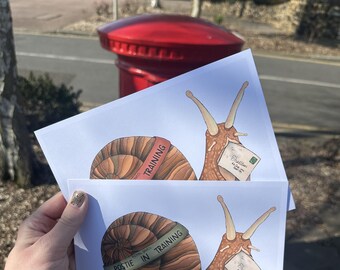 Op de afbeelding: Twee geïllustreerde postkaarten met een slak met een groen-wit lint om de schelp. De bovenste kaart zegt "Training" en de onderste kaart zegt "Postie in Training". Beide slakken dragen een brief met de tekst "Elvesboro" erop.