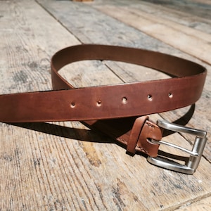 May include: A brown leather belt with a silver buckle. The belt is lying on a wooden surface.