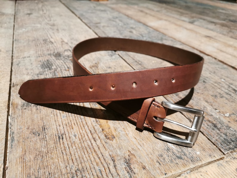 May include: A brown leather belt with a silver buckle. The belt is lying on a wooden surface.