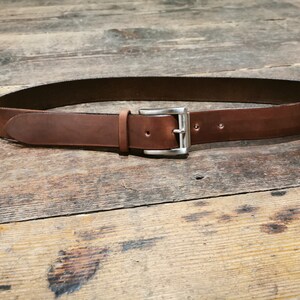 May include: A brown leather belt with a silver buckle. The belt is lying flat on a wooden surface.