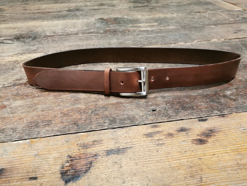 May include: A brown leather belt with a silver buckle. The belt is lying flat on a wooden surface.