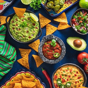 Può includere: Uno sfondo blu con una varietà di salse e snack. Ci sono ciotole di guacamole, salsa e salsa al formaggio. Ci sono anche patatine tortilla, jalapenos e fette di avocado.
