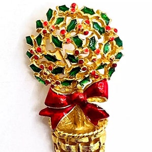 Könnte beinhalten: Eine goldfarbene Brosche mit einem Stechpalmenkranz mit roten Beeren in einem goldenen Korb mit einer roten Schleife.