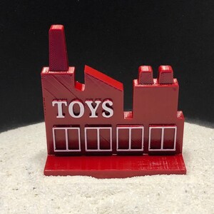 以下が含まれることがあります： 白い窓と正面に「TOYS」と書かれた赤いおもちゃ工場の建物です。