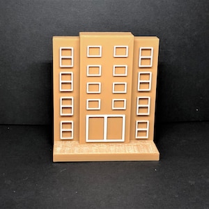 Può includere: Un modello di edificio di colore beige con finestre e una porta bianche. L'edificio ha cinque piani ed è fatto di plastica.