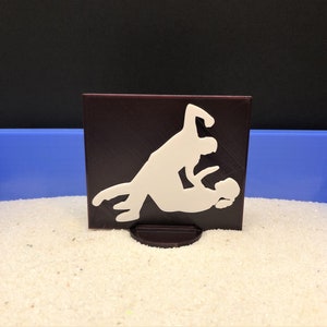 May include: A white silhouette of two people dancing on a dark brown square base. The base is sitting on a white sandy surface.