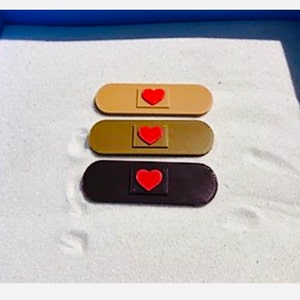 Op de afbeelding: Drie pleister-vormige objecten met rode harten op een wit zand oppervlak. De pleisters zijn beige, bruin en zwart.
