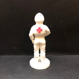 May include: White figurine of a person wearing a white uniform with a red cross on the back. The figurine is standing on a white base.