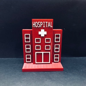May include: A red plastic model of a hospital building with white windows and a white cross on the front. The building says "HOSPITAL" on the top.