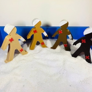 Op de afbeelding: Vier speelgoedfiguren van mensen met verbanden om hun hoofd en armen, elk met een rood kruis op hun romp, bevinden zich in een blauwe zandbak gevuld met wit zand.
