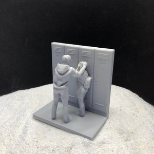 May include: A 3D printed figurine of two people standing in front of a row of lockers. The person on the left is holding the person on the right's arm. The figurine is painted in a light gray color.
