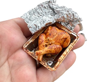 May include: A miniature roasted chicken in a foil tray. The chicken is a golden brown colour, with realistic details. The tray has a metallic finish, and the foil is crumpled. A small, detailed food item, likely for a dollhouse or display.