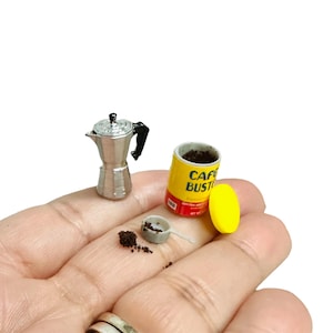 May include: Miniature coffee-making set featuring a silver stovetop espresso maker, a yellow can of "Cafe Busto" coffee, a small scoop, and a pile of coffee grounds. The set is held in a hand, suggesting a small scale.