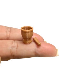May include: A miniature light brown mortar and pestle set. The mortar is a small, hollow, cup-shaped vessel with a flared base, and the pestle is a tiny, slender, cylindrical tool with a rounded end, both crafted in a smooth, solid light brown material.
