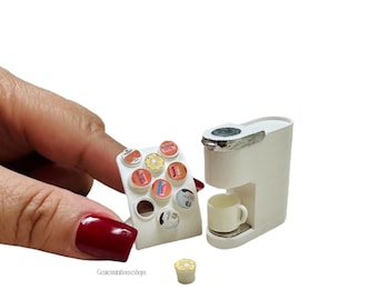 May include: Miniature white coffee maker with a silver accent, a small white mug, and a selection of coffee pods. The coffee maker is next to a holder with various coffee pod designs. The image is on a white background.
