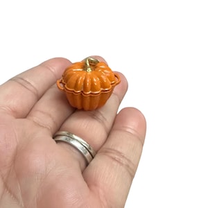 May include: A small orange pumpkin-shaped casserole dish with a gold handle. The dish is about the size of a person's hand.