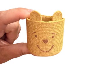 May include: A small, cylindrical, mustard-yellow woven basket featuring two rounded ears at the top. The exterior surface displays a stitched brown facial design consisting of two simple eyes, a small nose, and a curved line representing a smile.