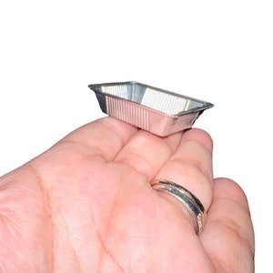 May include: A small, rectangular, silver-coloured aluminium foil baking pan with a ribbed exterior. The pan is empty and appears unused. The pan is held in a person's hand.