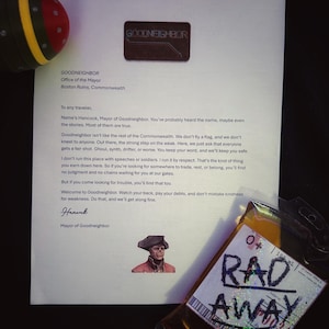 May include: A printed letter from Goodneighbor's Mayor, Hancock, with the text "Goodneighbor" at the top. A brown patch with the same text is above the letter. A bag of "Rad Away" is visible, along with a red and yellow object.