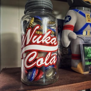 Pode incluir: Um frasco de vidro transparente cheio de tampinhas de garrafa, com a inscrição "Nuka Cola" em vermelho e branco. O frasco tem uma tampa prateada e está sobre uma prateleira de madeira. Uma figura de astronauta é visível no fundo.