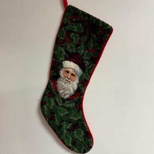 May include: A needlepoint Christmas stocking featuring a central portrait of Santa Claus with a white beard and red hat, surrounded by a dense pattern of green holly leaves and red berries. The stocking has a red fabric trim and a red hanging loop.