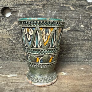 May include: An ornate, handcrafted ceramic vase with a detailed metalwork design. The vase features a vibrant, patterned exterior with blue, yellow, and white accents. The metalwork is silver-toned, adding a touch of elegance to the piece. The vase has a small, round base.