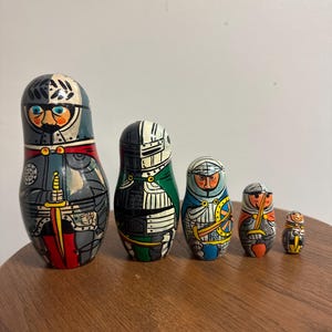 Könnte beinhalten: Ein Set aus fünf ineinander stapelbaren Holzfiguren, die als Ritter in Rüstung bemalt sind. Jede Figur zeigt einen individuellen Helm, einen Gesichtsausdruck und detaillierte Rüstungselemente in Grau, Rot, Grün, Blau und Gelb.