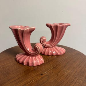 May include: A pair of pink ceramic candle holders featuring a cornucopia-inspired shape with a flared, scalloped base and a curved, scroll-like design. The glossy finish highlights the vertical ridges along the body of each individual piece.