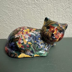May include: A ceramic cat figurine in a resting pose, adorned with a colorful floral pattern. The cat's body is covered in a mosaic of red, yellow, blue, and green flowers. The figurine is a decorative item.