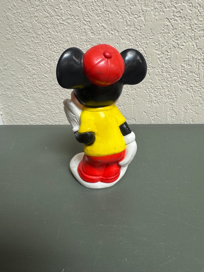 May include: A vintage Mickey Mouse figurine, viewed from the back. The figure wears a red cap, yellow shirt, red shorts, and white gloves. The ears are black, and the base is white. The figurine is a classic Disney collectible.