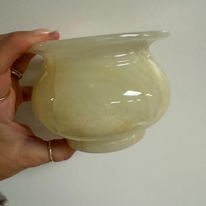 Può includere: Una ciotola di onice traslucida giallo pallido con un corpo arrotondato e un bordo svasato. La ciotola ha una superficie liscia e lucida e una piccola base circolare. L'onice presenta sottili variazioni di colore. La ciotola è tenuta da una mano.