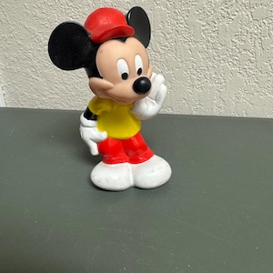 May include: A Mickey Mouse figurine in a classic pose, wearing a red cap, yellow shirt, red pants, and white gloves and shoes. The iconic character has black ears and a black nose. The figurine is standing on a dark surface.