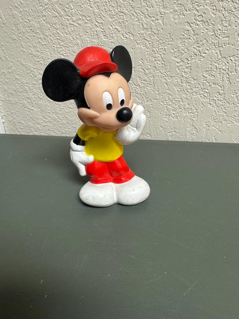 May include: A Mickey Mouse figurine in a classic pose, wearing a red cap, yellow shirt, red pants, and white gloves and shoes. The iconic character has black ears and a black nose. The figurine is standing on a dark surface.