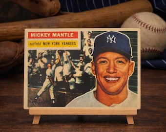 May include: Vintage baseball card of Mickey Mantle, outfielder for the New York Yankees, displayed on a small wooden easel. The card features a smiling portrait of Mantle in a Yankees cap, with a team photo in the background.