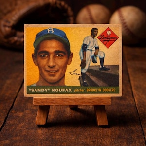 Könnte beinhalten: Vintage Baseballkarte mit Sandy Koufax, Pitcher der Brooklyn Dodgers. Die Karte zeigt ein Porträt und eine Actionaufnahme von Koufax, vor einem gelb-grünen Hintergrund. Die Karte wird auf einer kleinen Holzstaffelei präsentiert.