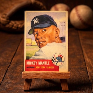 May include: A vintage Mickey Mantle baseball card on a small wooden easel. The card shows Mantle in a New York Yankees cap. Text on the card reads "Mickey Mantle, outfielder, New York Yankees". The card is displayed on a wooden surface.