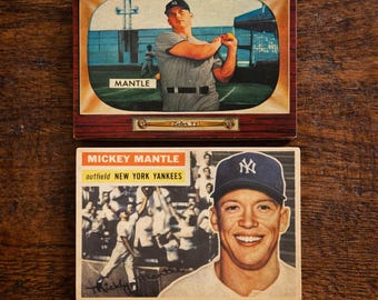 May include: Two vintage baseball cards featuring Mickey Mantle. One card shows Mantle in a pinstripe uniform on a TV screen, the other shows a smiling portrait of Mantle in a Yankees cap. The cards are on a wooden surface.