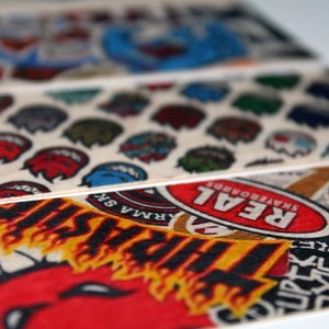 May include: Close-up of several skateboard grip tapes with various colorful graphics. The designs include cartoon faces, logos, and text, with a focus on red, blue, and black colors. The tapes are arranged in a layered fashion.