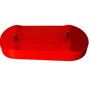 Puede incluir: Un objeto rectangular rojo brillante con extremos redondeados. La superficie tiene un patrón texturizado con líneas paralelas. Dos pasadores metálicos son visibles en la parte superior. Probablemente es un componente de un objeto más grande.