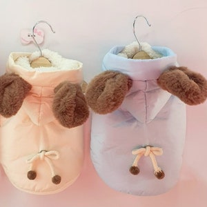 Peut inclure: Deux manteaux pour chiens, un rose clair et un bleu clair, chacun avec des accents en forme d'oreilles marron. Les manteaux ont un cordon de serrage en bas et une doublure blanche duveteuse autour de la capuche. Ils sont suspendus à des cintres métalliques.