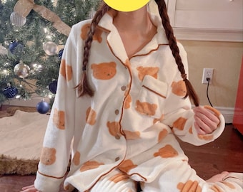 May include: White pyjamas with a brown bear print. The pyjamas have long sleeves and long trousers. The pyjamas have a button-up front and a pocket on the left side.
